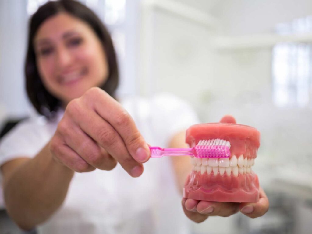 Dentista demonstrando como limpar prótese protocolo em casa com escova e modelo de dentes na Clínica R7 Implantes no Catete RJ.