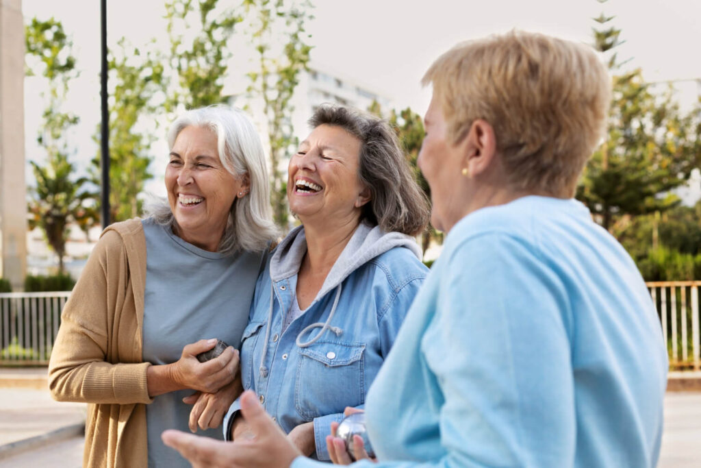Três amigas seniores rindo ao ar livre. Na R7 Implantes no Catete - RJ, o metal do implante dentário não fica visível no sorriso.